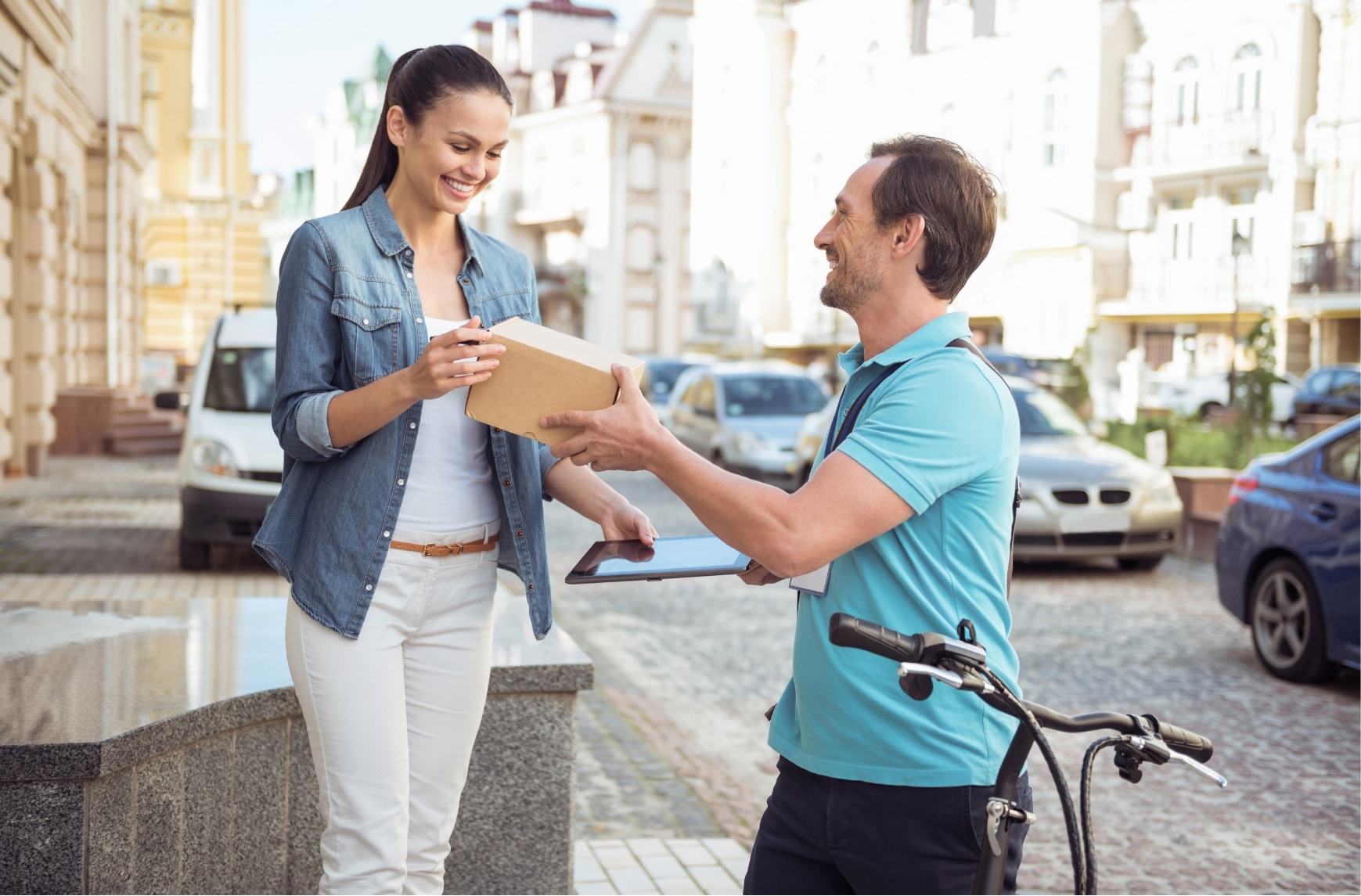 A man on a bicycle deliver a package to a woman in an urban area.